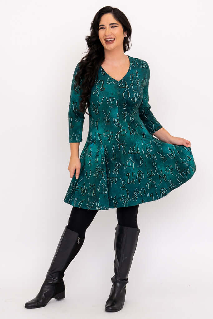 Model smiling in teal tunic with abstract design, holding skirt with hand, black leggings, and boots