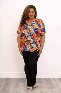 Woman posing in a vivid floral button-up blouse and black pants, hands crossed at waist