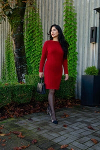 Woman in a red cable knit sweater dress, standing outdoors next to green ivy, holding a black handbag