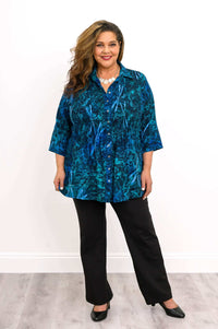 Woman in blue smocked shirt and black pants standing against a white background