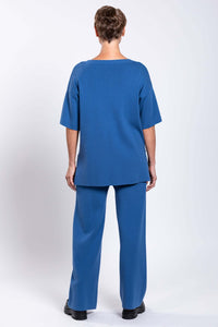 Back view of woman wearing blue top and blue knit pants and black shoes against a white background