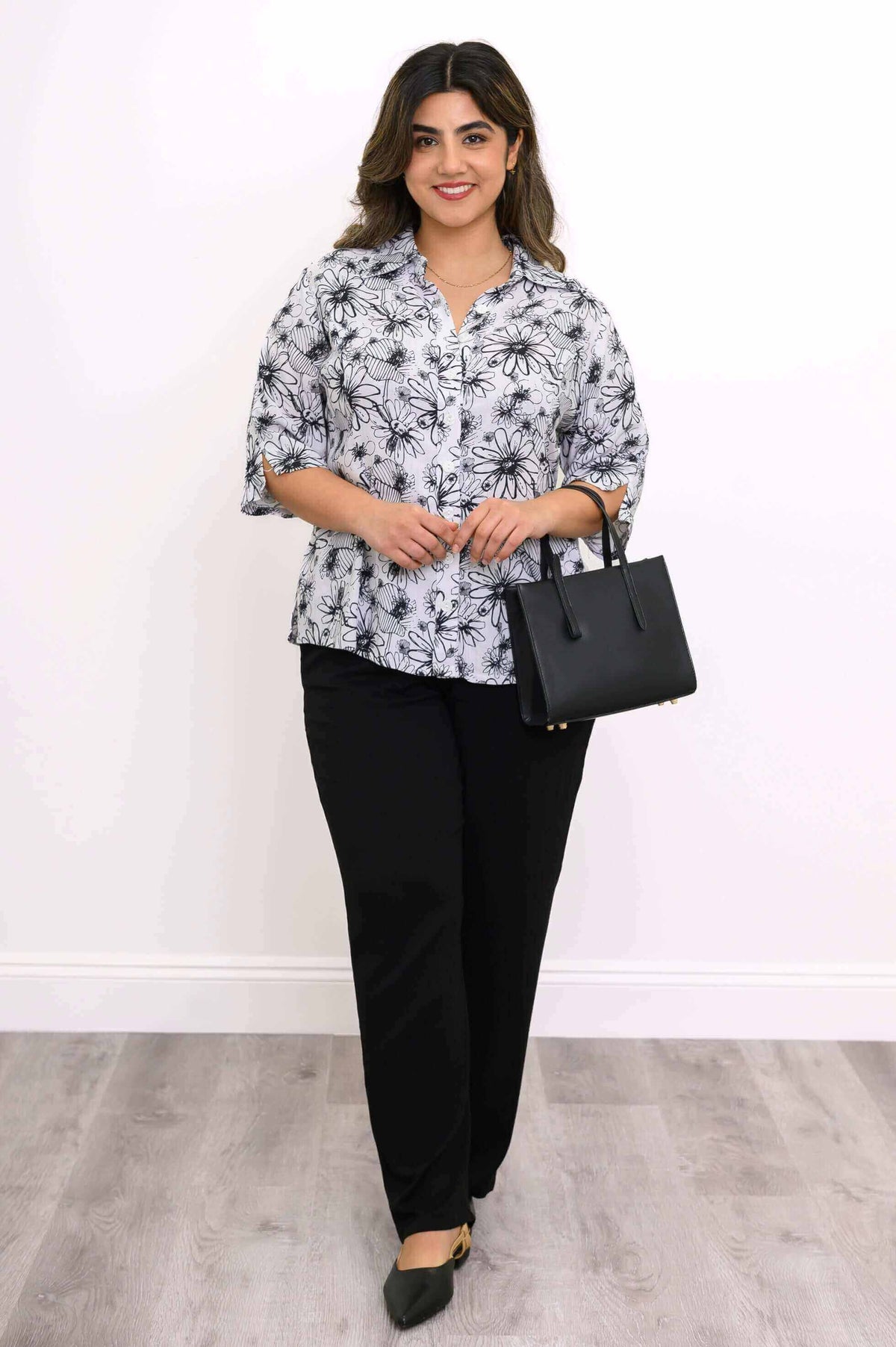 Model holding a black handbag, wearing black and white floral blouse and black pants