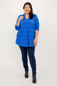 Model with hand on shoulder, wearing a blue patterned tunic, navy pants, and boots
