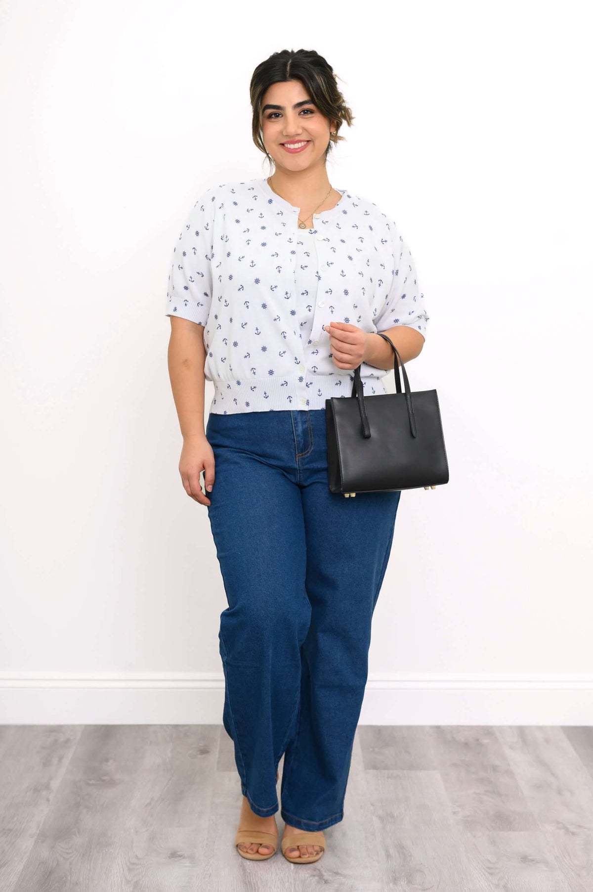 Woman standing in a white nautical print cardigan, blue jeans, tan sandals, and holding a black handbag