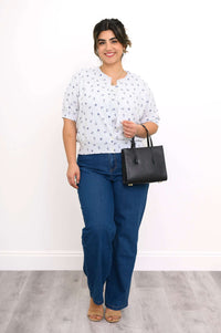 Woman standing in a white nautical print cardigan, blue jeans, tan sandals, and holding a black handbag