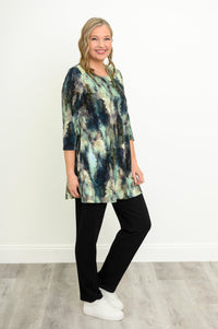 Woman in a blue-green watercolor forest print A-line tunic, black trousers, and white sneakers against a white wall.