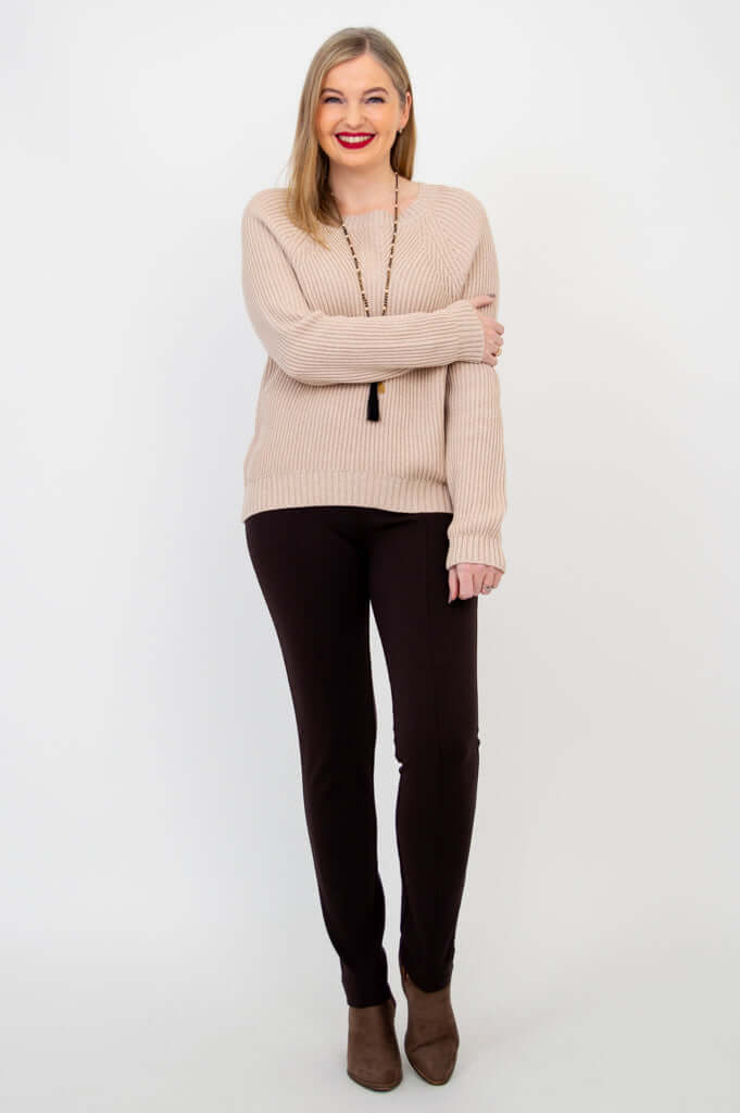 Woman in beige knit sweater and brown pants posing with arm across body