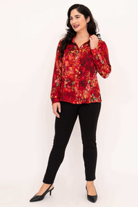 Woman posing in red floral blouse with hand near collar and black pants