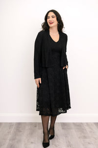 Woman smiling and posing in lace overlay dress styled with a black shrug and black heels