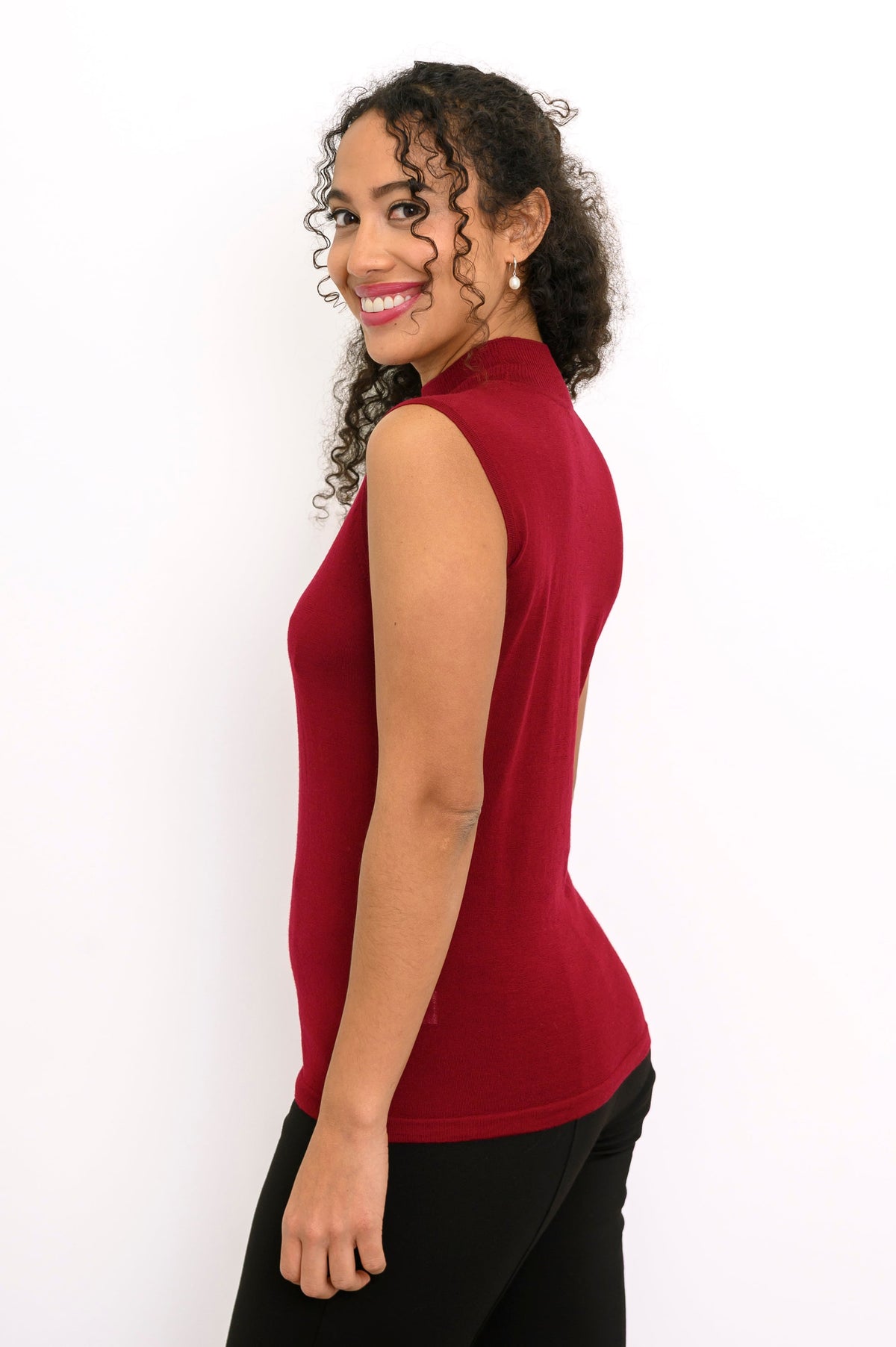 Side profile view of a woman wearing a sleeveless burgundy merino wool top styled with black pants.