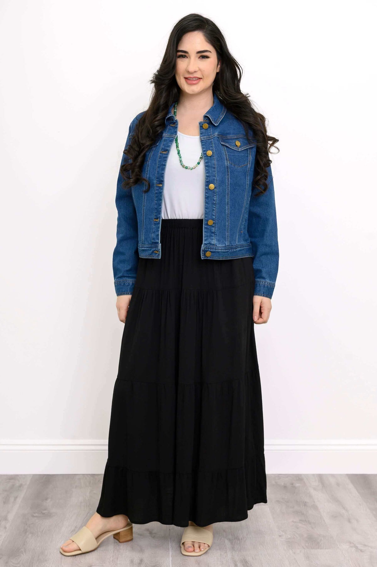 Woman in black tiered maxi skirt with white top, denim jacket, and green necklace