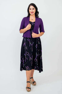 Woman in black dragonfly print sleeveless dress with purple shrug, hand on chest