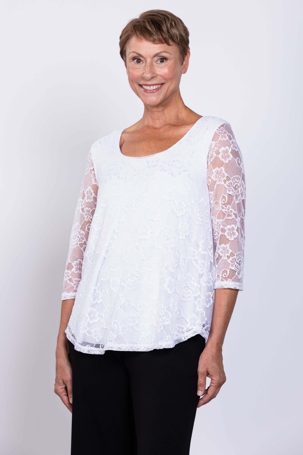 Side-front view of a woman smiling in a white lace overlay 3/4 sleeve top and black pants