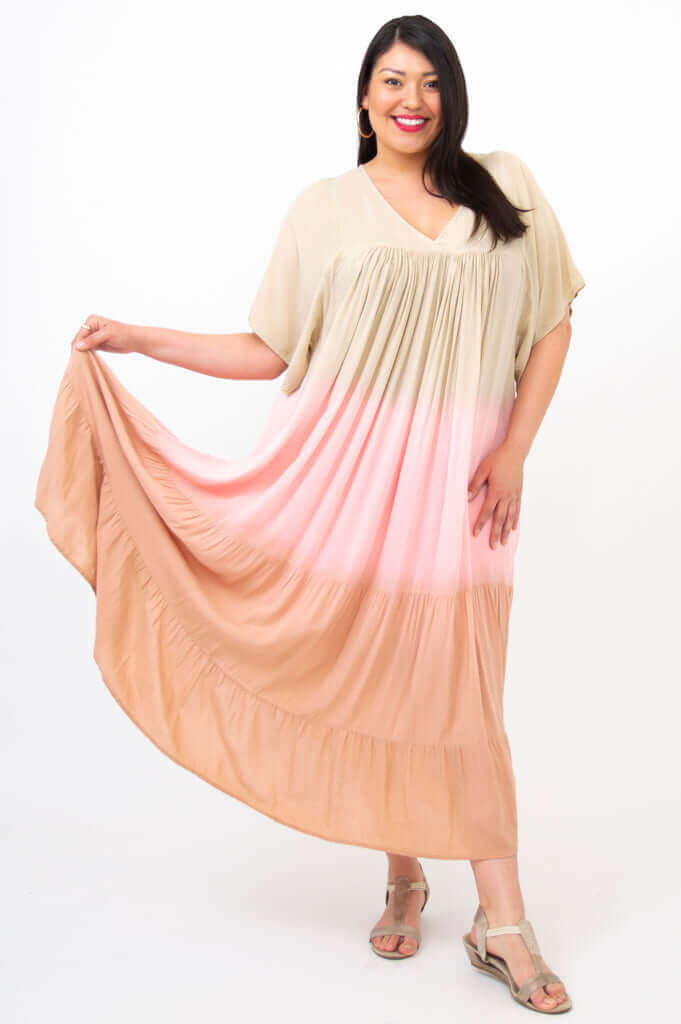 Woman posing in a beige, pink, and peach tiered ombre maxi dress, holding the skirt to the side