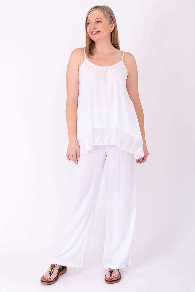 Side-facing woman in a white lace-trimmed tank top and white pants, smiling