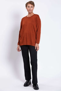 Woman in rust red sweater standing angled to the side, wearing black pants and boots