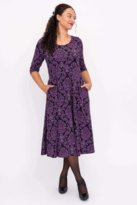 Side angled front view of model in a purple damask midi dress with pockets