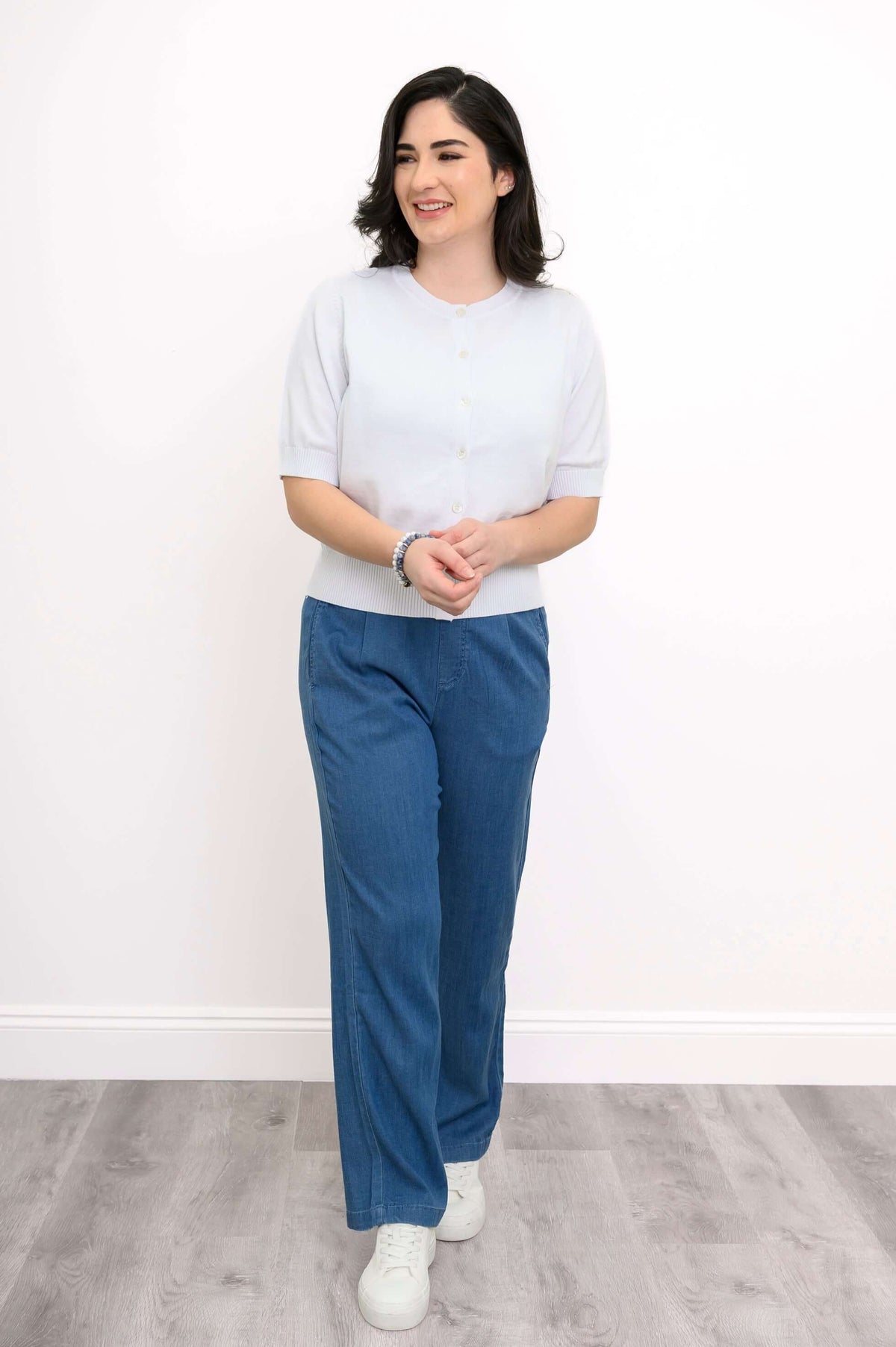 Side-angled pose of woman wearing a white cardigan with blue pants and white sneakers