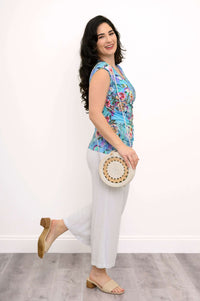 Woman posing sideways in a blue floral faux wrap tank top and light pants, holding a round bag