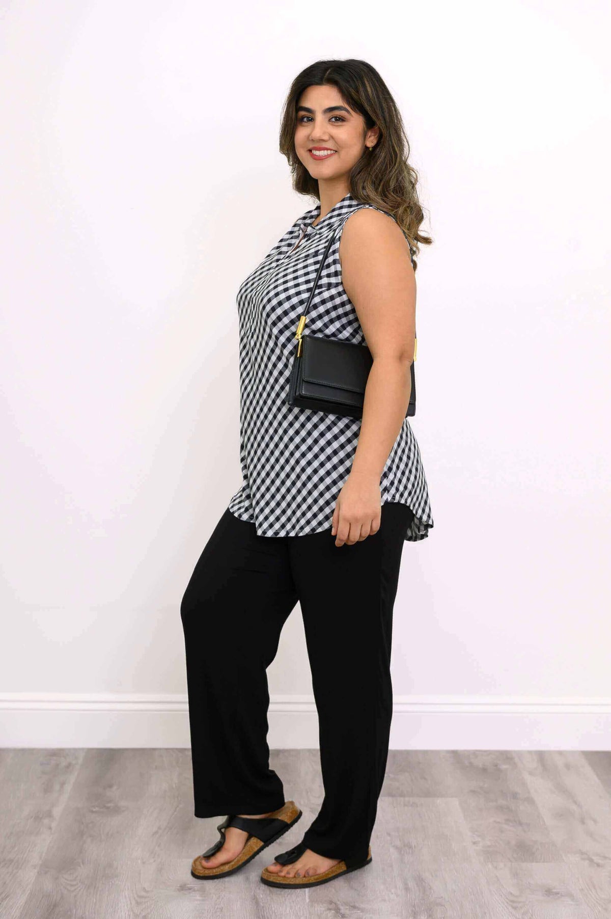Side view of a woman in a gingham tank top, black pants, and sandals with a black handbag