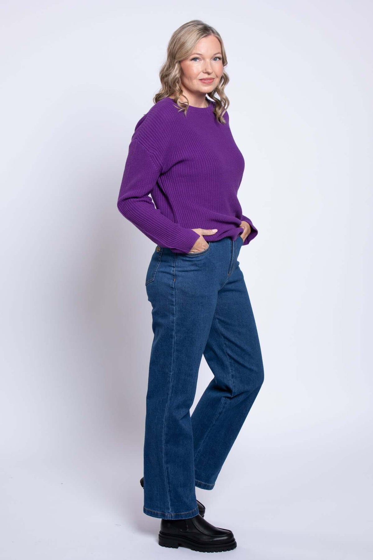 Side view of a woman in blue straight leg jeans and purple sweater, hands in pockets.
