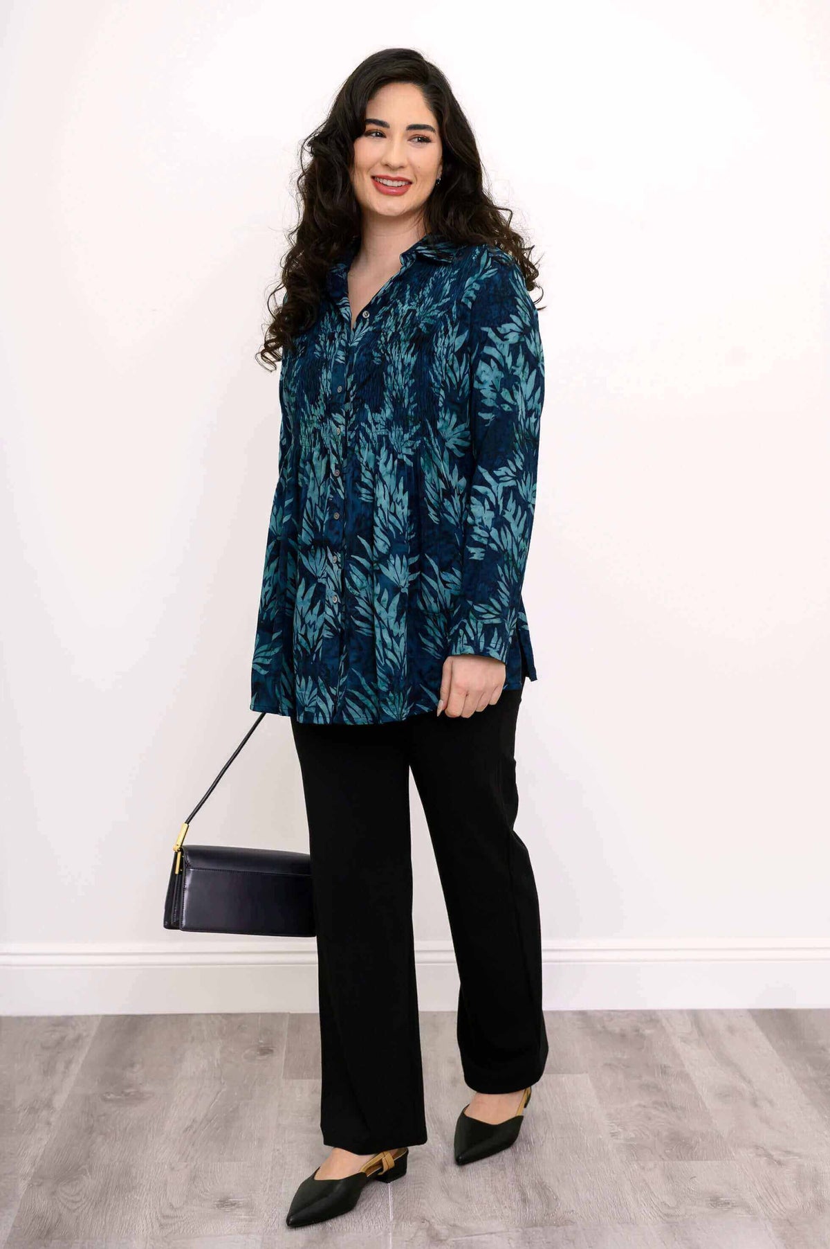 Side view of model wearing blue smocked tunic, black pants, and carrying a purse