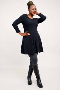 Model standing sideways with hands in pockets, wearing a black tunic dress