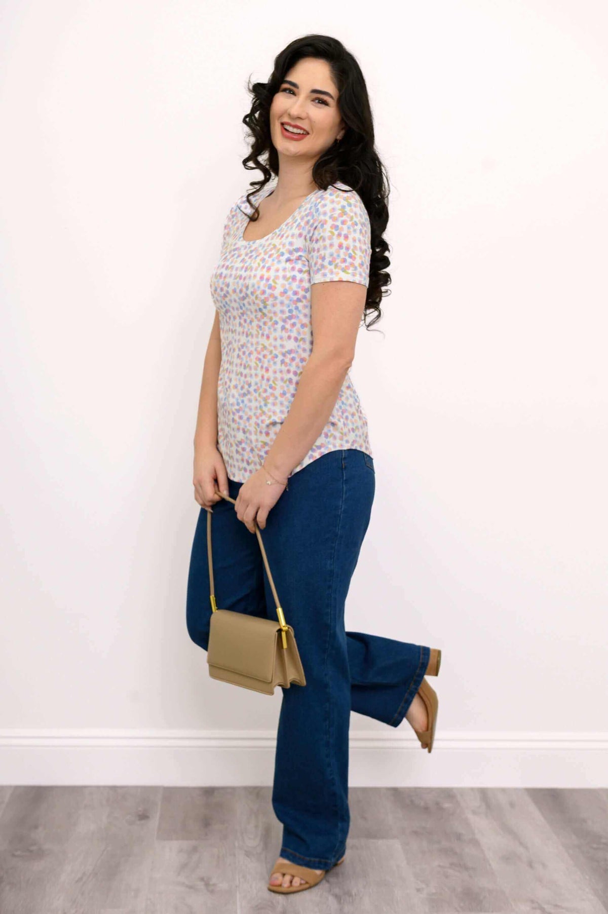 Woman in pastel confetti tee, jeans, and tan bag, smiling and standing sideways