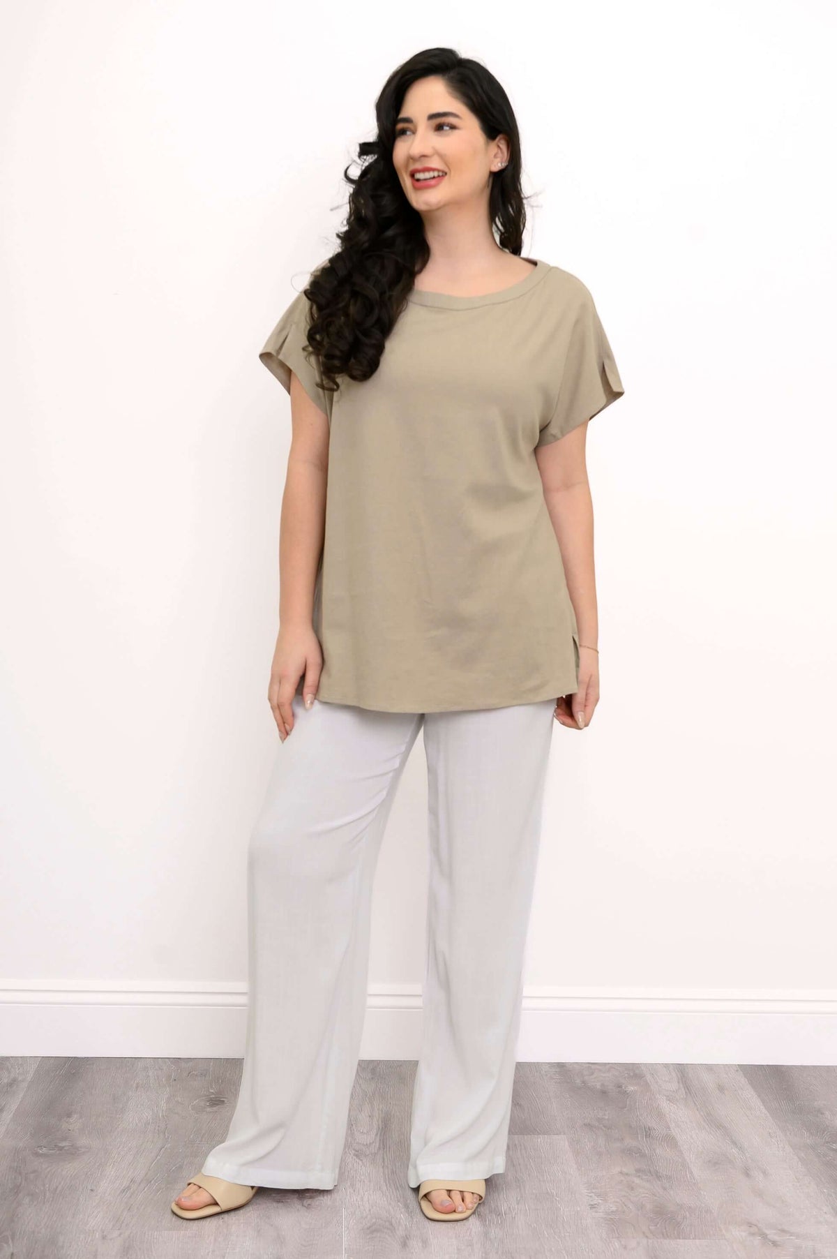Woman in taupe tee with cap sleeves and white pants, smiling and looking right