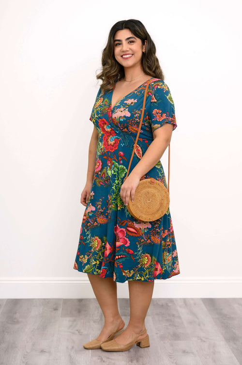 Model in a teal floral V-neck dress, showing the side and holding a round rattan bag
