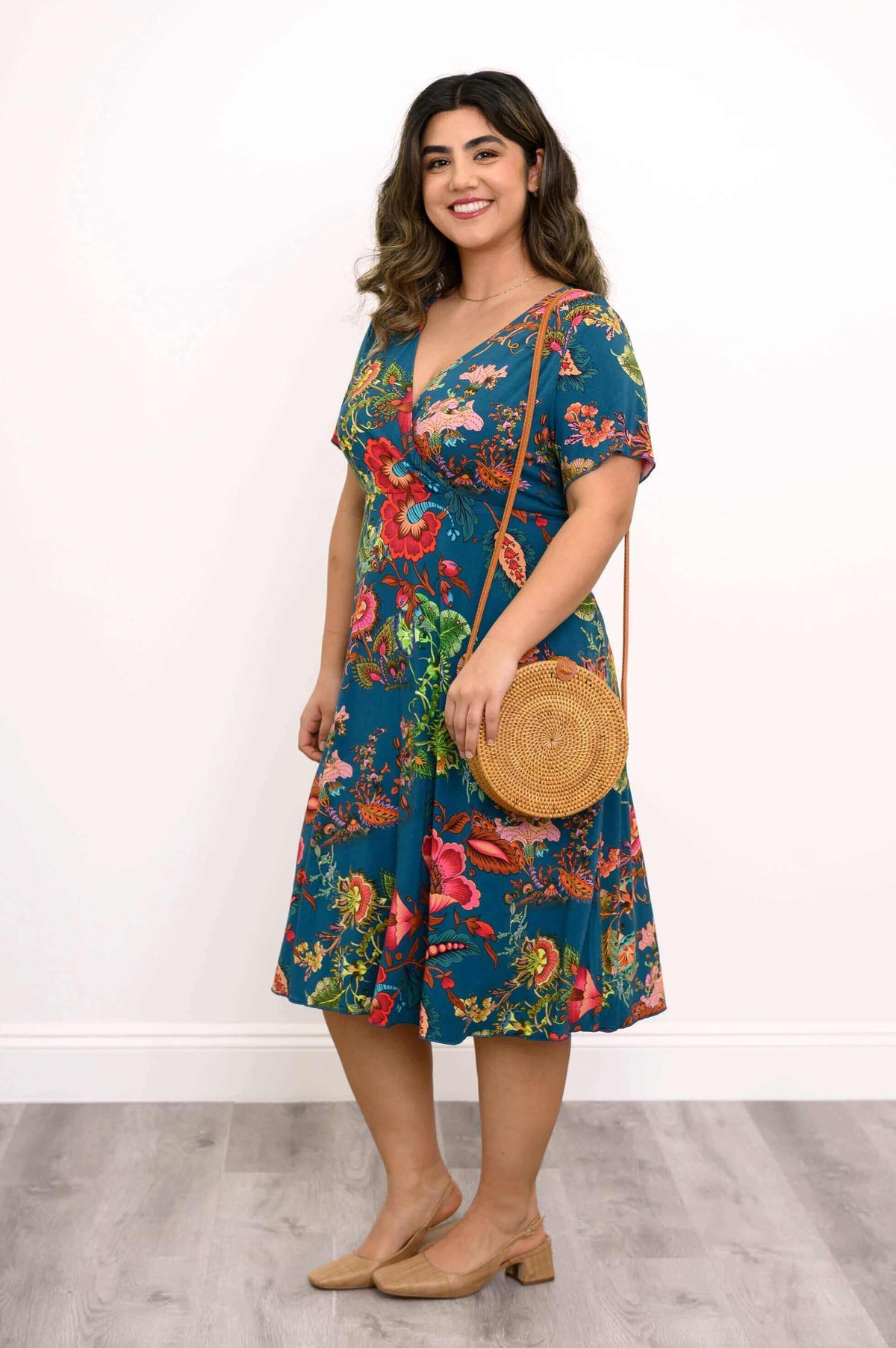 Side view of a woman in a teal floral bamboo wrap dress holding a round straw bag
