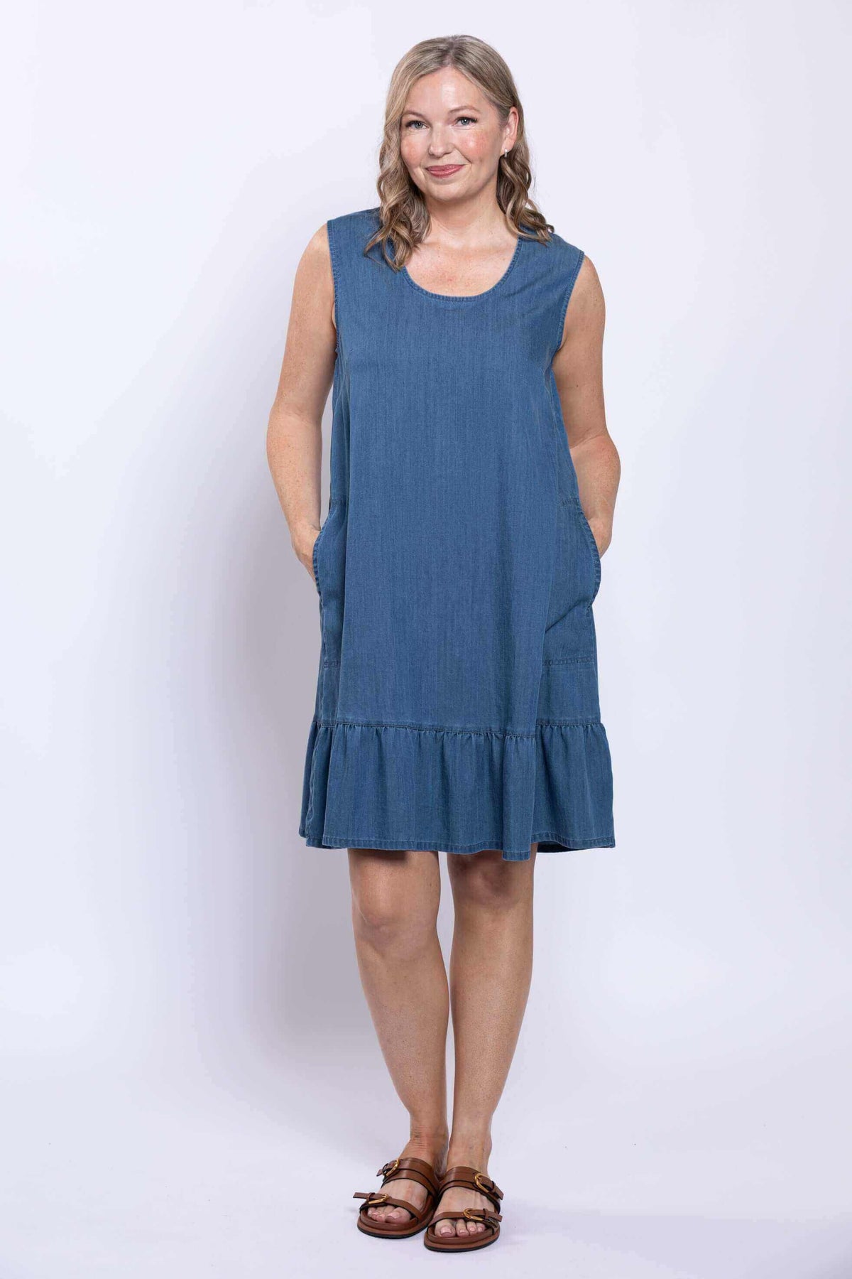 Woman wearing a sleeveless denim blue dress with ruffle hem and side pockets, front view