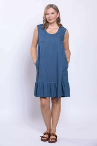 Woman wearing a sleeveless denim blue dress with ruffle hem and side pockets, front view