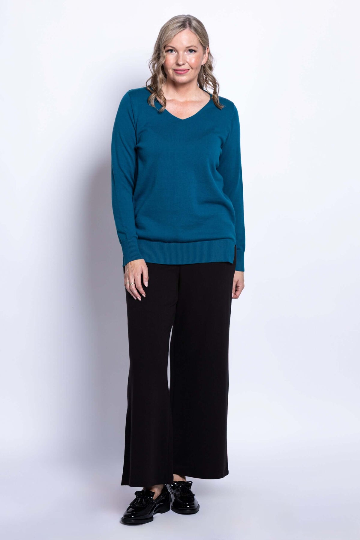 Slightly angled front view of woman in teal V-neck cotton sweater and black pants
