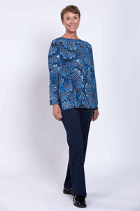 Full body view of a smiling woman wearing a blue botanical print tunic top with navy pants.