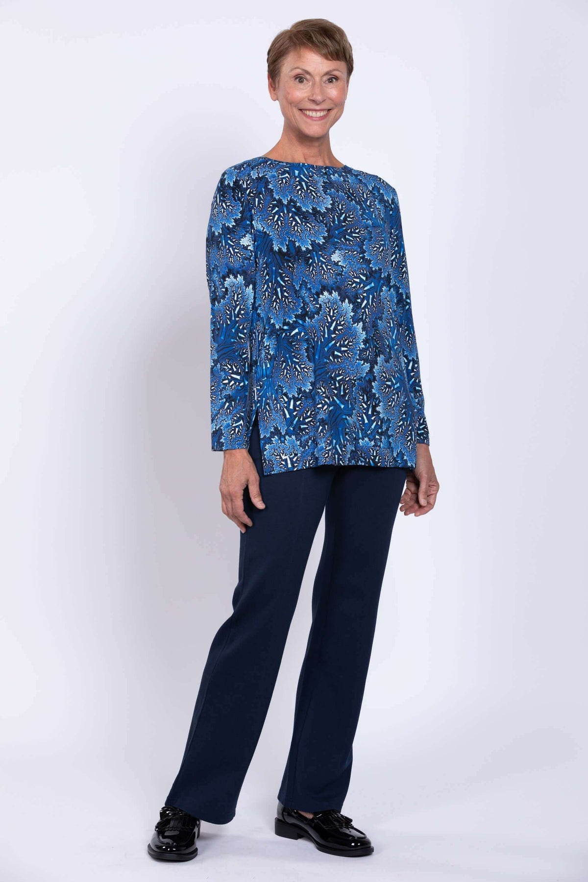 Angled full body view of a smiling woman wearing a blue botanical print top with navy pants.