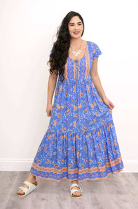 Woman smiling in a blue boho maxi dress with floral and peach details, holding the skirt