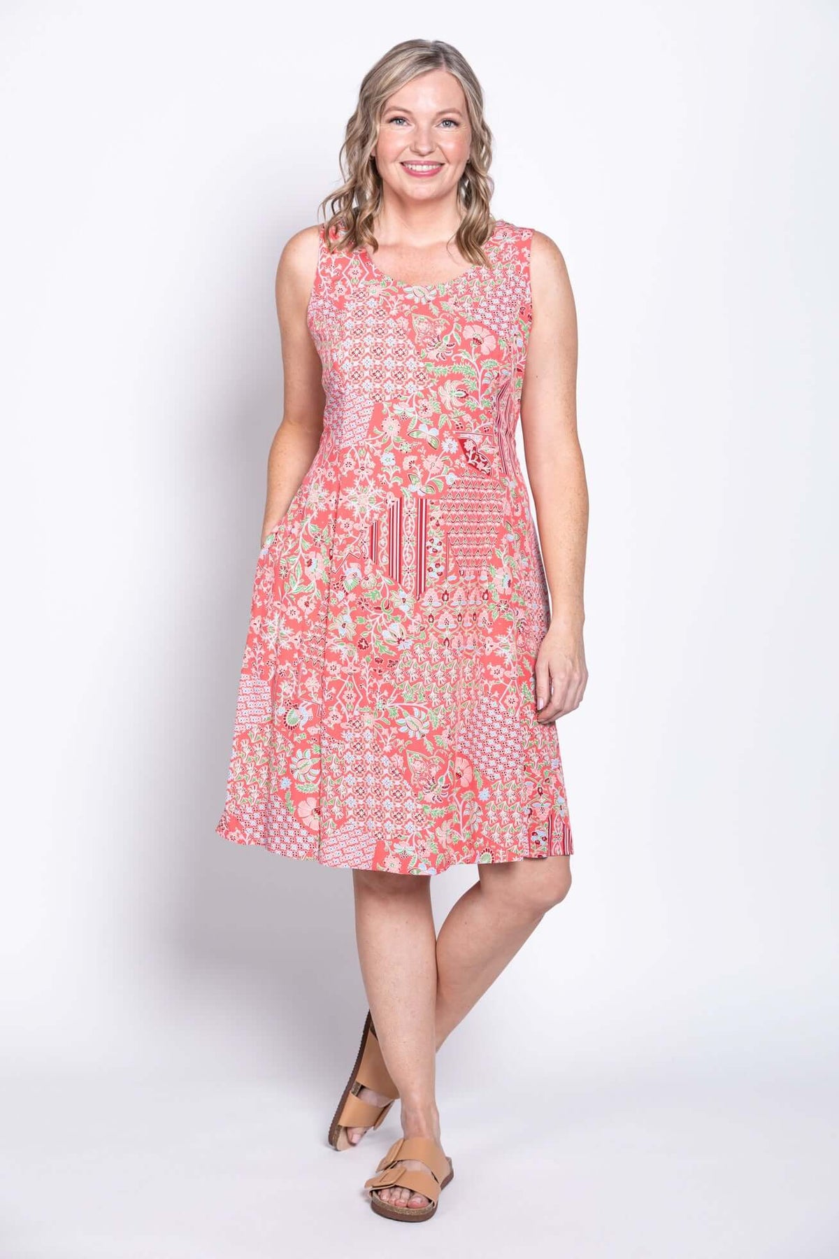 Woman smiling in a coral floral sleeveless dress, hands in pockets, front view