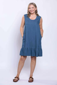 Woman in denim blue sleeveless dress, smiling with hands in pockets, front view
