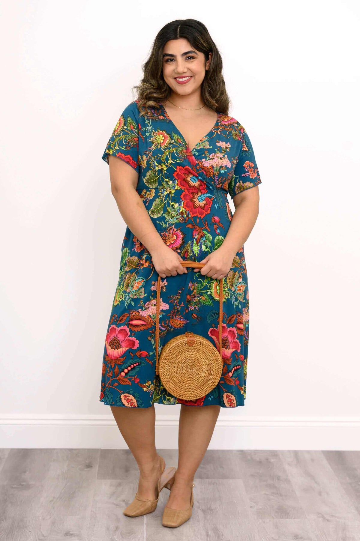 Woman smiling and holding a round straw bag, wearing a teal floral bamboo wrap dress