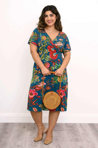 Woman smiling and holding a round straw bag, wearing a teal floral bamboo wrap dress