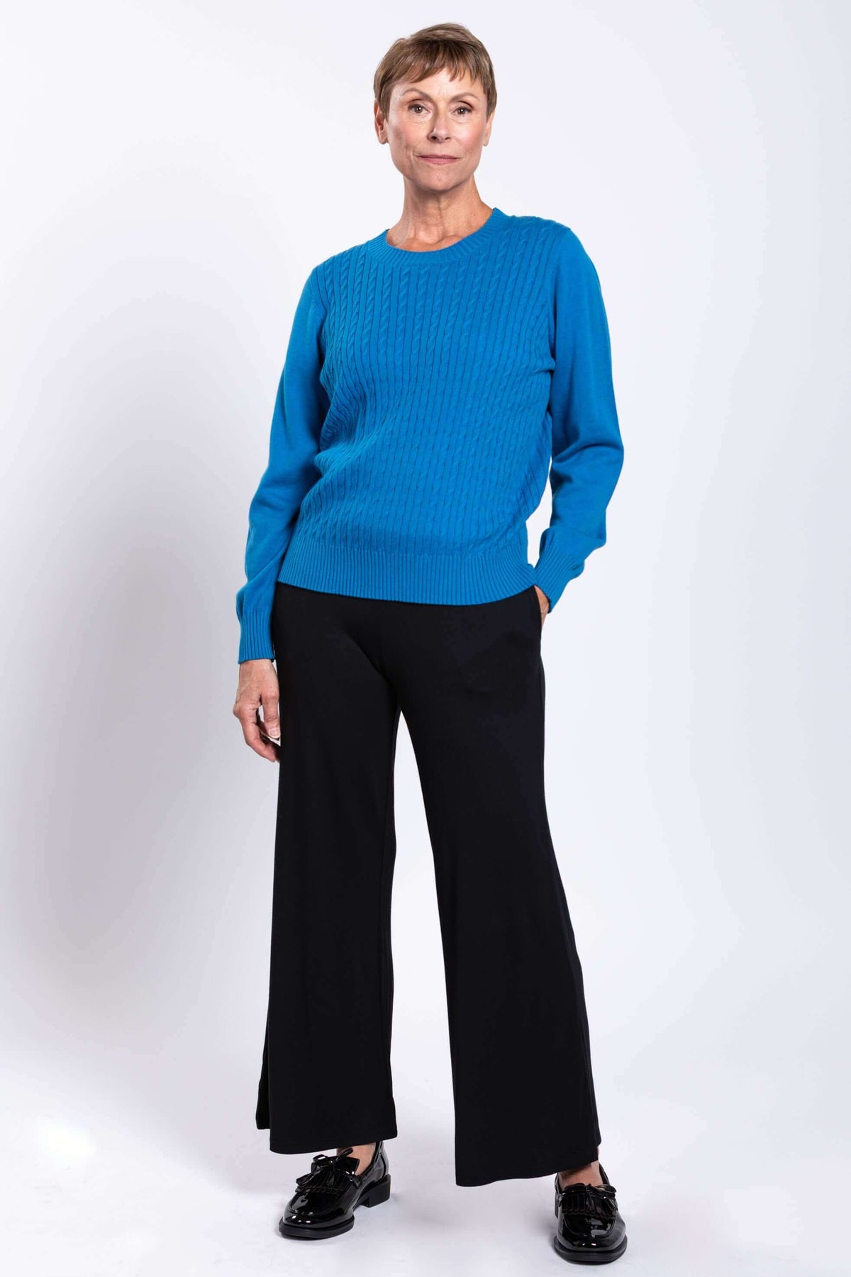 Woman standing with one hand in pocket, modeling a blue cable knit sweater and black wide-leg pants