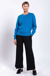 Woman standing with one hand in pocket, modeling a blue cable knit sweater and black wide-leg pants