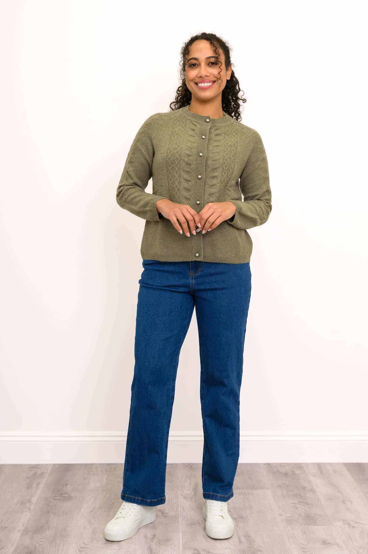 Woman standing in green cable knit cardigan with hands together, blue jeans, white sneakers
