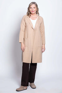 Woman standing in a beige open front cotton jacket, white shirt, and brown pants