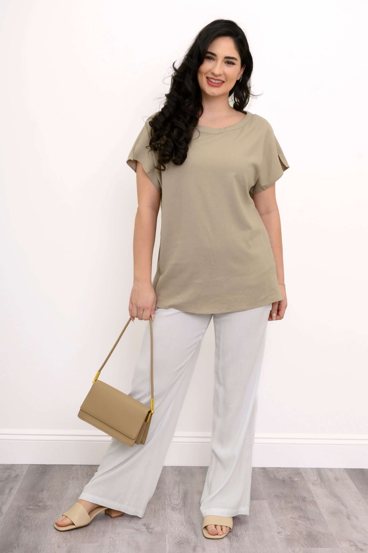 Woman smiling in taupe tee and white pants holding a beige shoulder bag