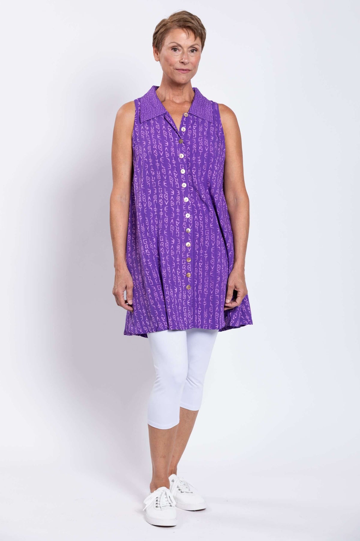 Three-quarter view of woman in violet patterned button up tank top over white leggings