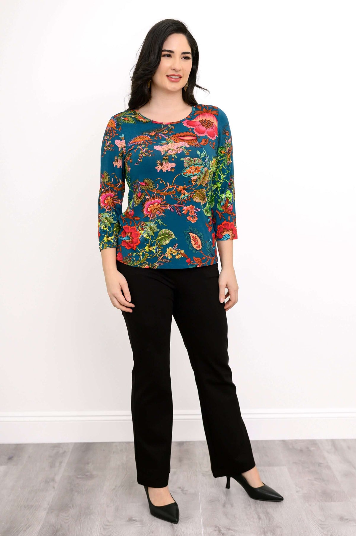 Woman in floral teal top and black pants, three-quarter turn, hands at sides
