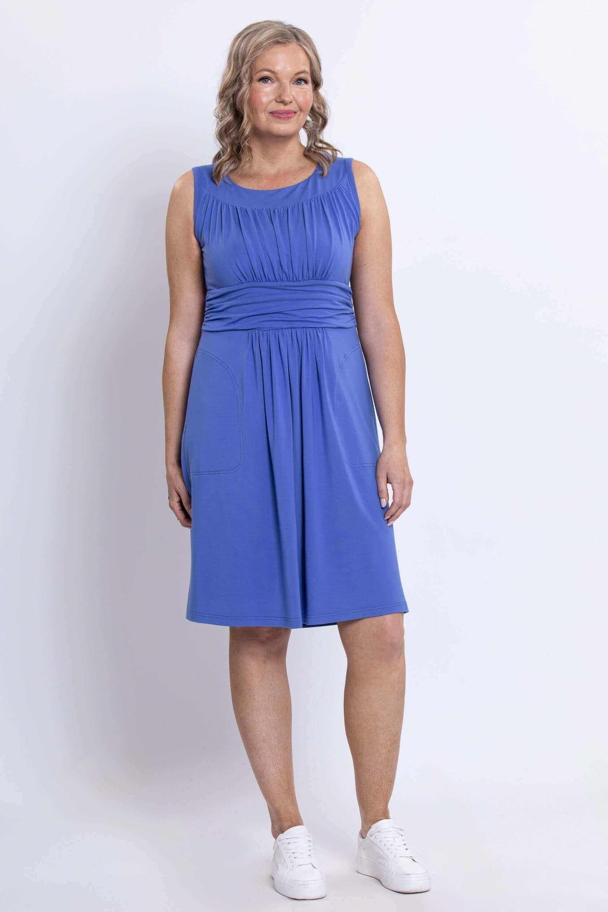Three-quarters front view of a woman in a sleeveless blue ruched dress with waistband