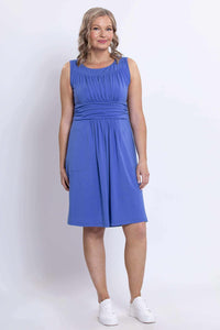 Three-quarters front view of a woman in a sleeveless blue ruched dress with waistband